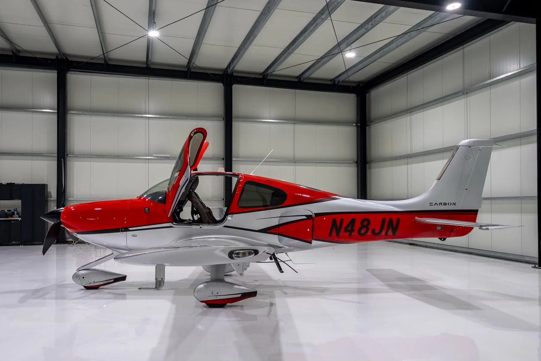 Side view of a red and silver Cirrus SR22T Carbon aircraft with registration N48JN and open doors, in a hangar.