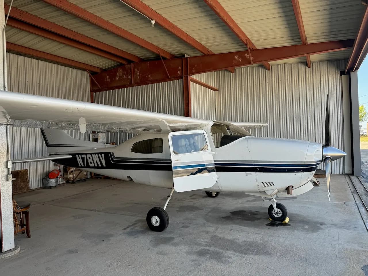 A side view of a white Cessna aircraft with blue and black stripes, registration N78WV, with its door open, parked in a hangar.