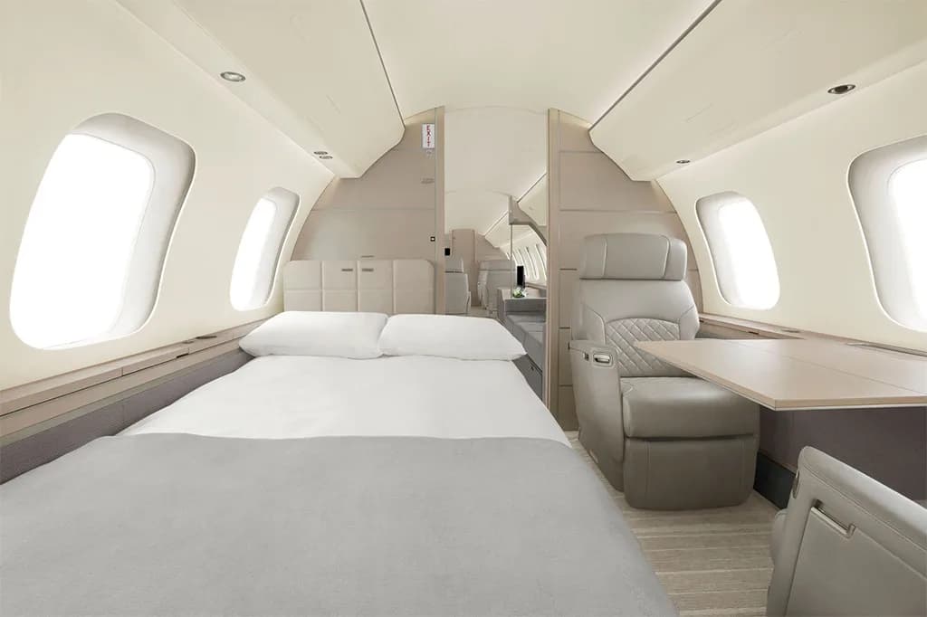 An aircraft cabin configured with a bed made with white sheets and a grey blanket, next to a light grey seat and table.