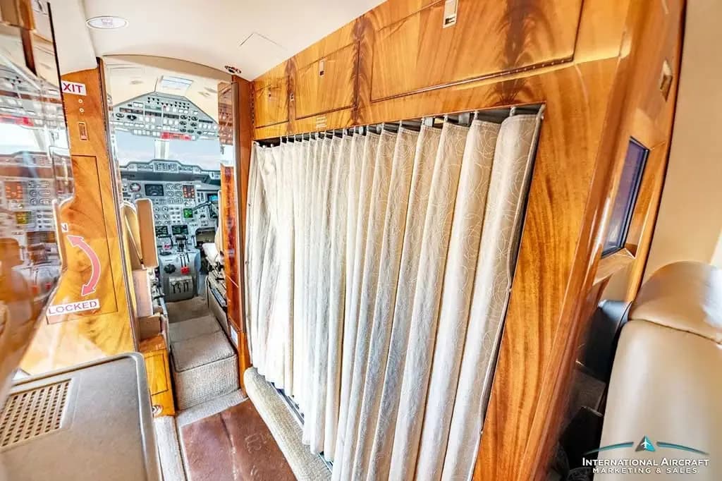 A beige curtain separates the luxurious wooden-paneled cabin from the cockpit of the private jet.