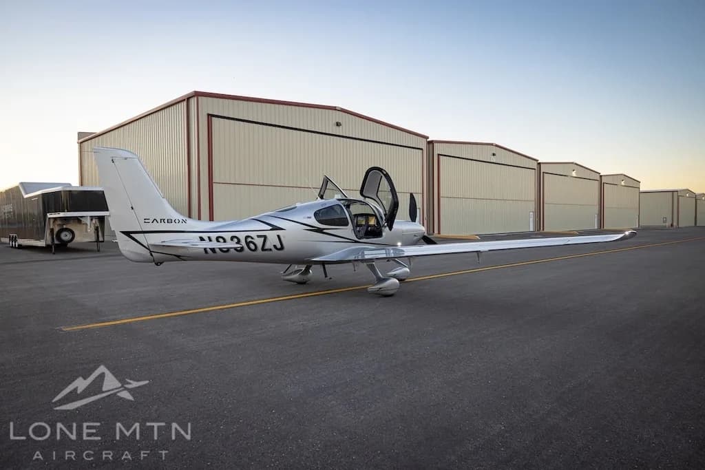 A silver Cirrus SR22T aircraft with registration N836ZJ is parked on an asphalt ramp with its doors open, in front of a row of hangars.