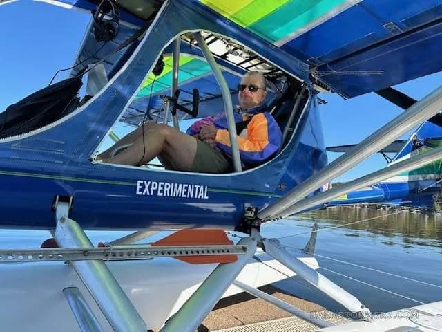 A man in sunglasses and a colorful jacket sits in the open cockpit of a blue experimental seaplane docked on the water.