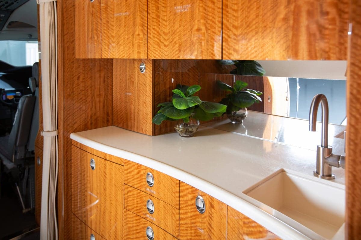 An aircraft galley area featuring wood cabinetry, a white countertop, a sink, and a view into the cockpit.