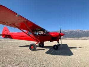 A red Cubcrafters XCub aircraft parked on a dirt airstrip with mountains in the background.