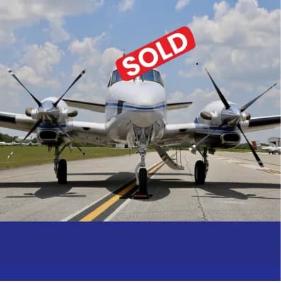 A white twin-engine turboprop aircraft with blue stripes is parked on an airport tarmac, viewed from the front.