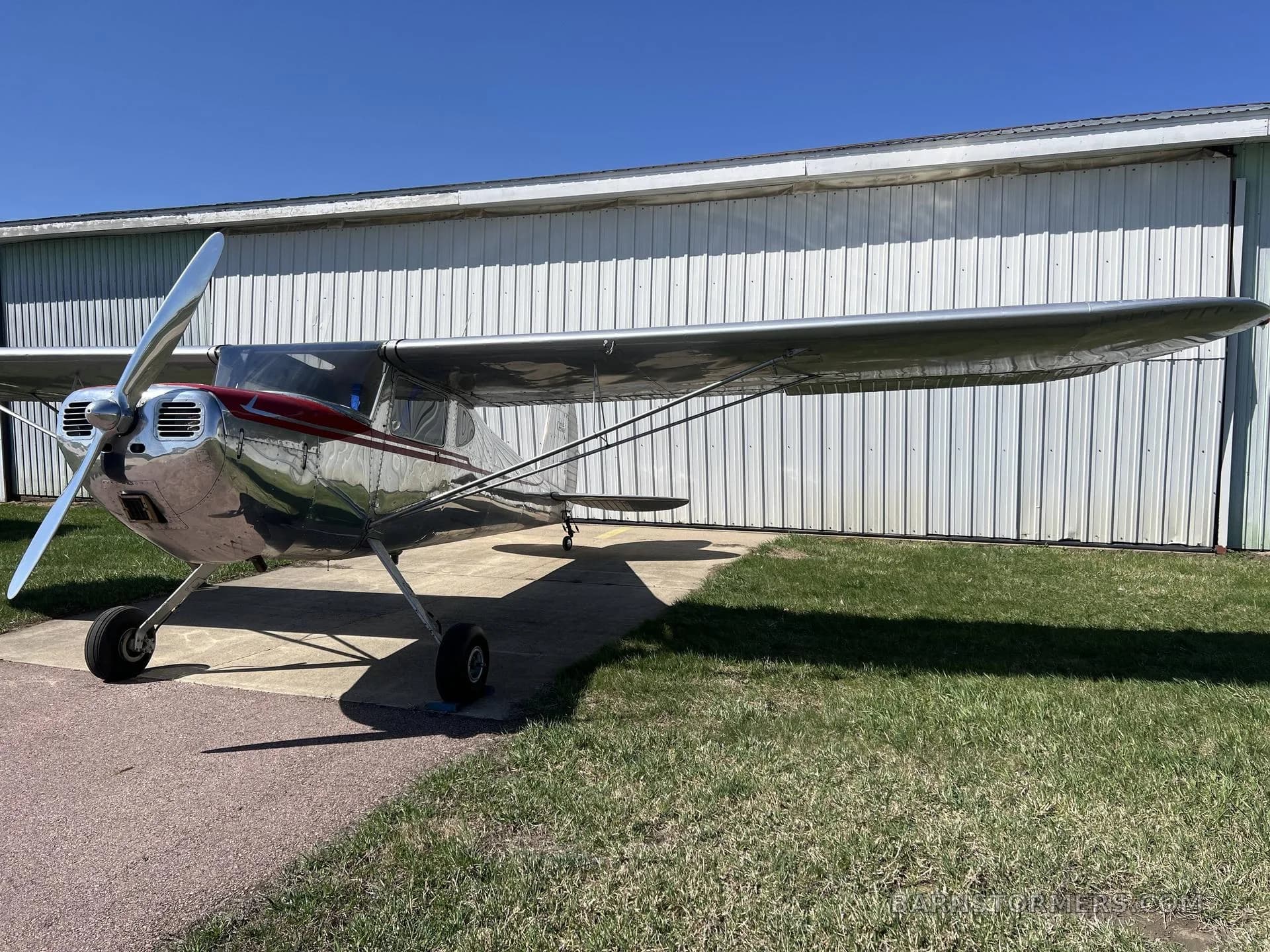 Front-quarter view of a silver Cessna 140 with a red cowling, parked on a concrete pad in front of a white hangar.