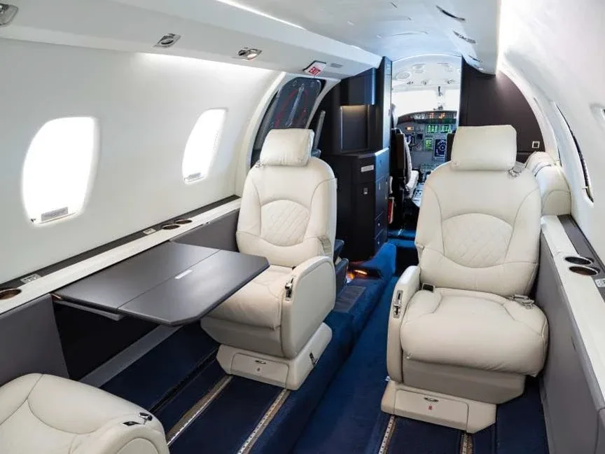 A view inside the business jet cabin, showing light beige leather seats, a deployed table, and a blue carpeted aisle.