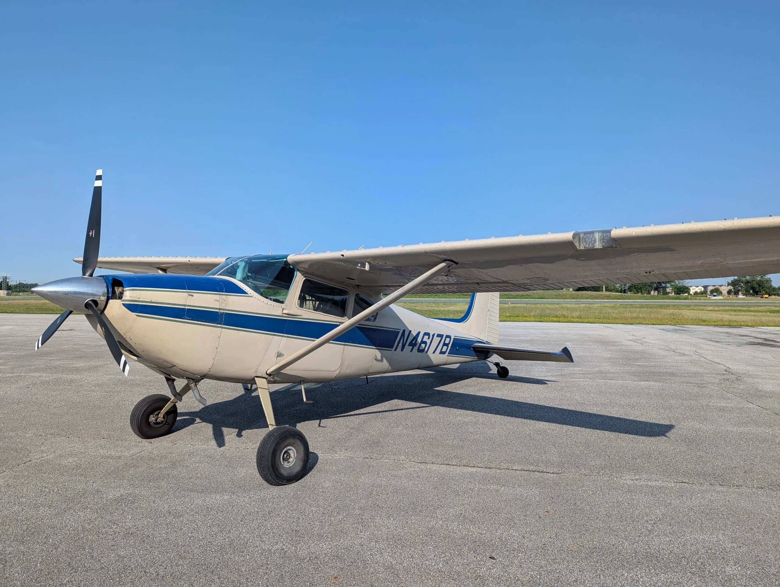 A beige and blue single-engine propeller aircraft, N4617B, is parked on an asphalt tarmac under a clear blue sky.