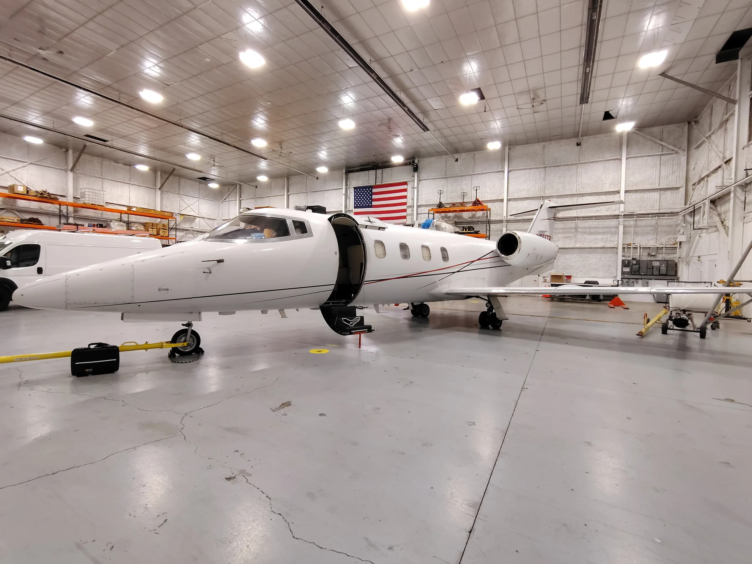 A white business jet with an open door is parked in a hangar with an American flag in the background.