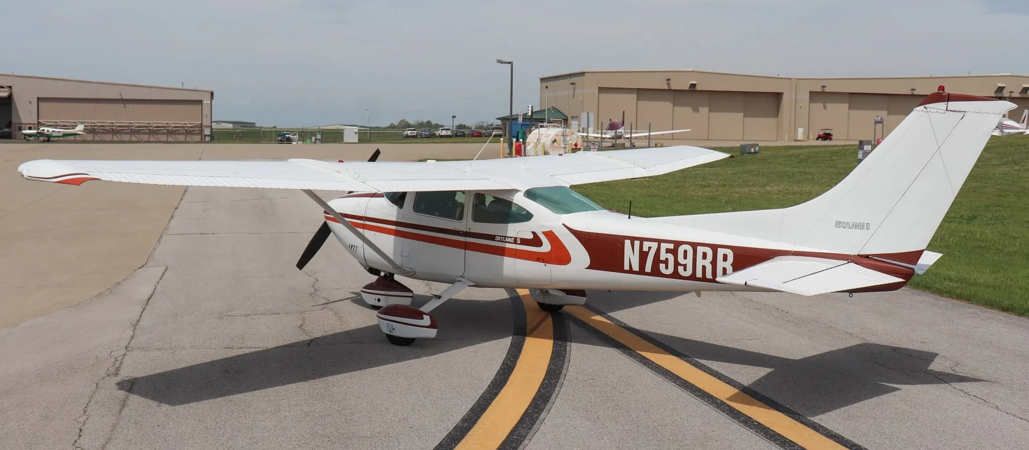 An aerial-like view of the Cessna Skylane S (N759RR) parked on an airfield with multiple hangars in the background.