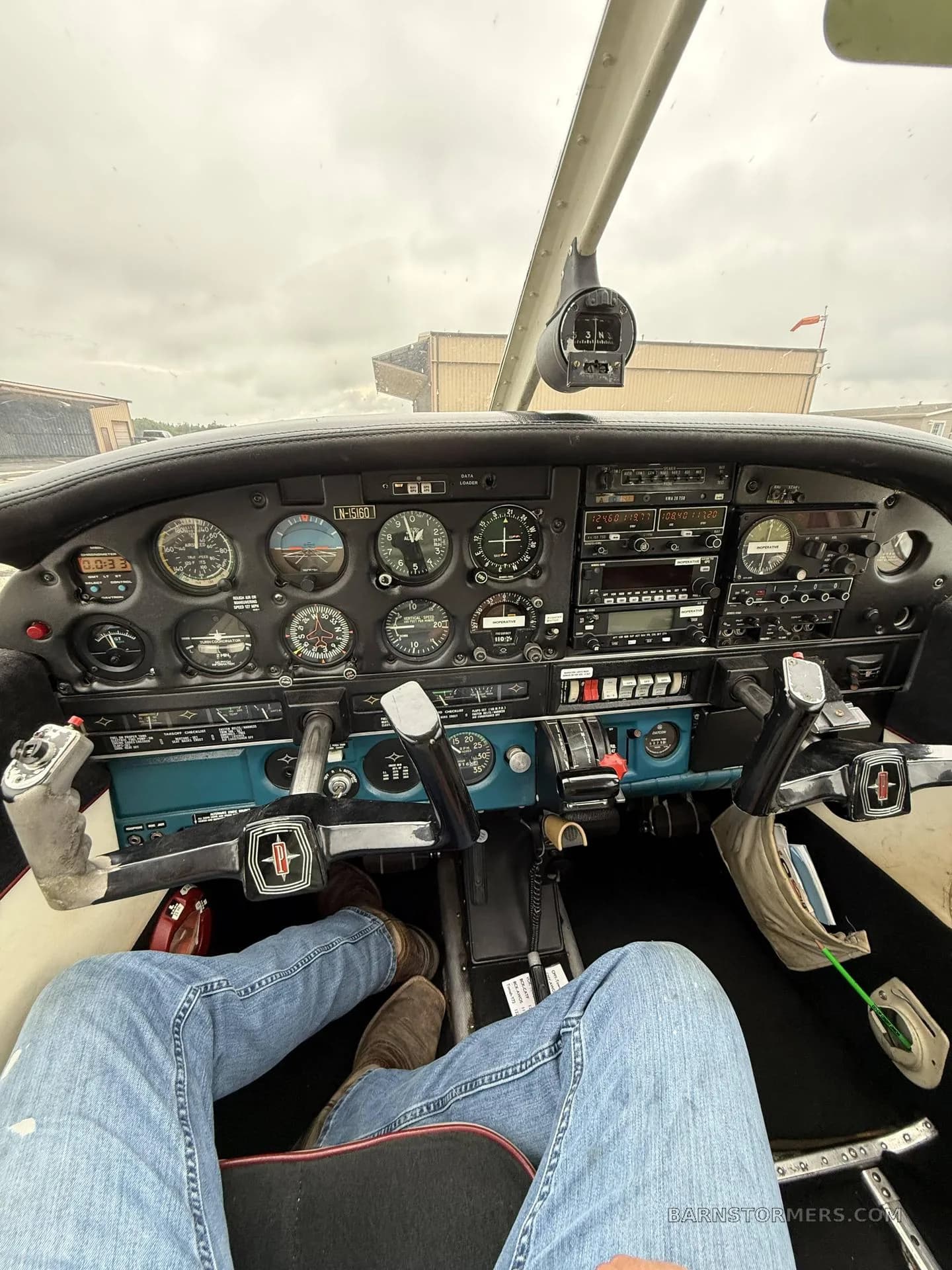 The cockpit of a light aircraft showing the instrument panel, controls, and a pilot's legs in jeans.