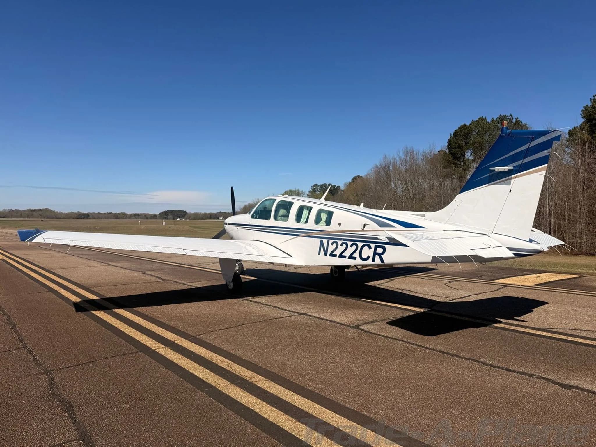 A white Beechcraft Bonanza N222CR with blue and gold stripes parked on an asphalt runway, viewed from the rear-left.