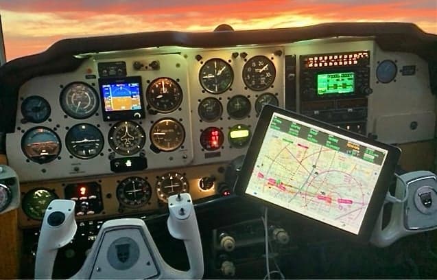 An aircraft cockpit showing the instrument panel, control yoke, and a tablet displaying a navigation map, with a sunset visible through the windshield.