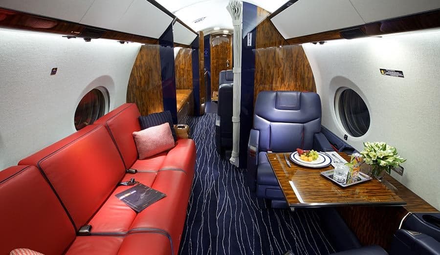 The luxurious cabin interior of a private jet, featuring a red leather sofa, blue leather seats, and dark wood accents.