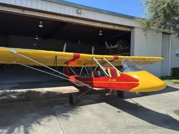 A red and yellow Air-Cam aircraft parked in front of a hangar.