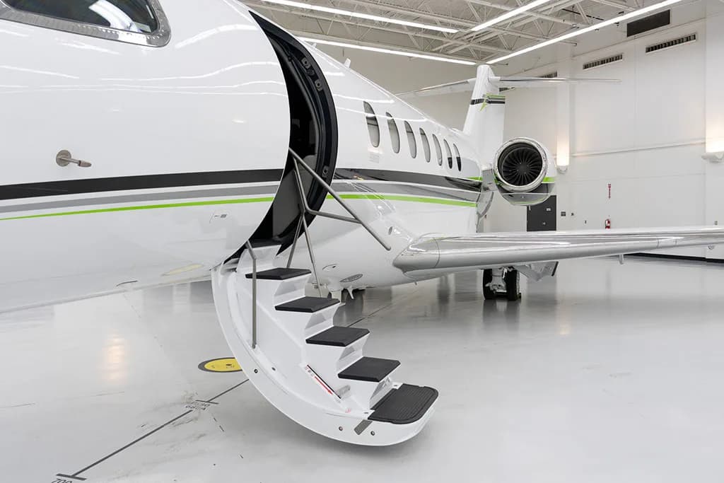 A close-up view of the open main door and extended stairs of a white business jet with green and black stripes in a hangar.