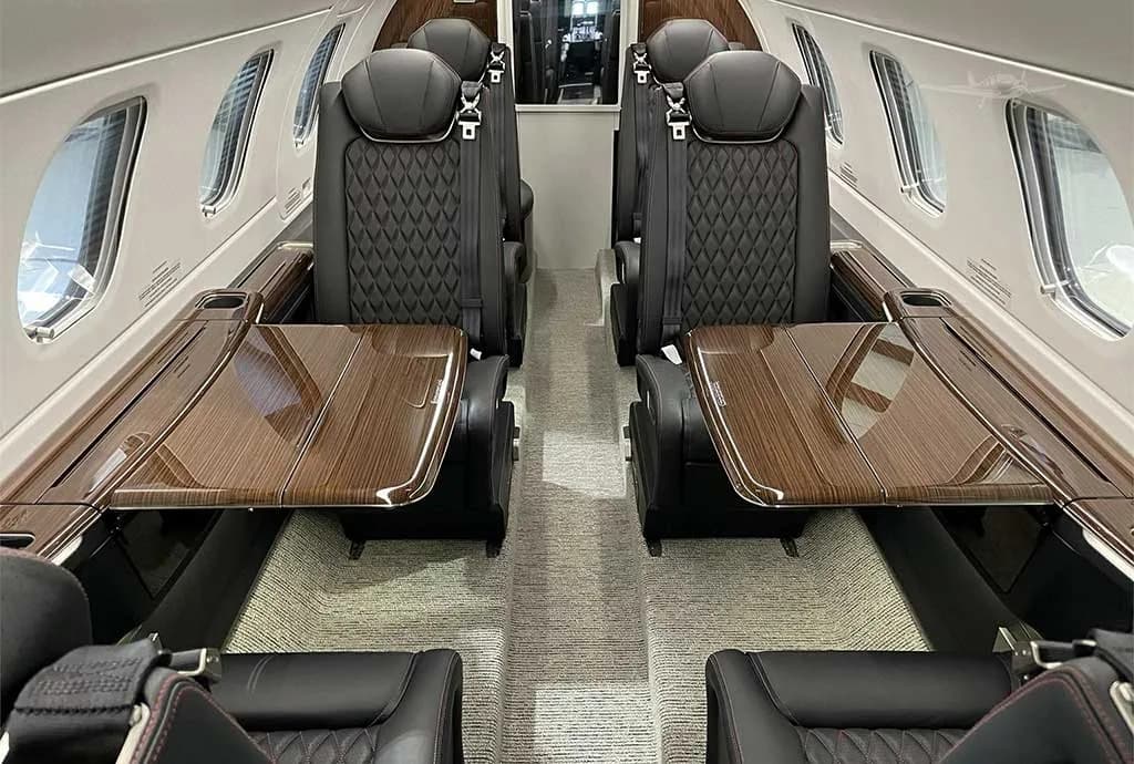 The luxurious interior cabin of a private jet with black quilted leather seats and wooden tables.