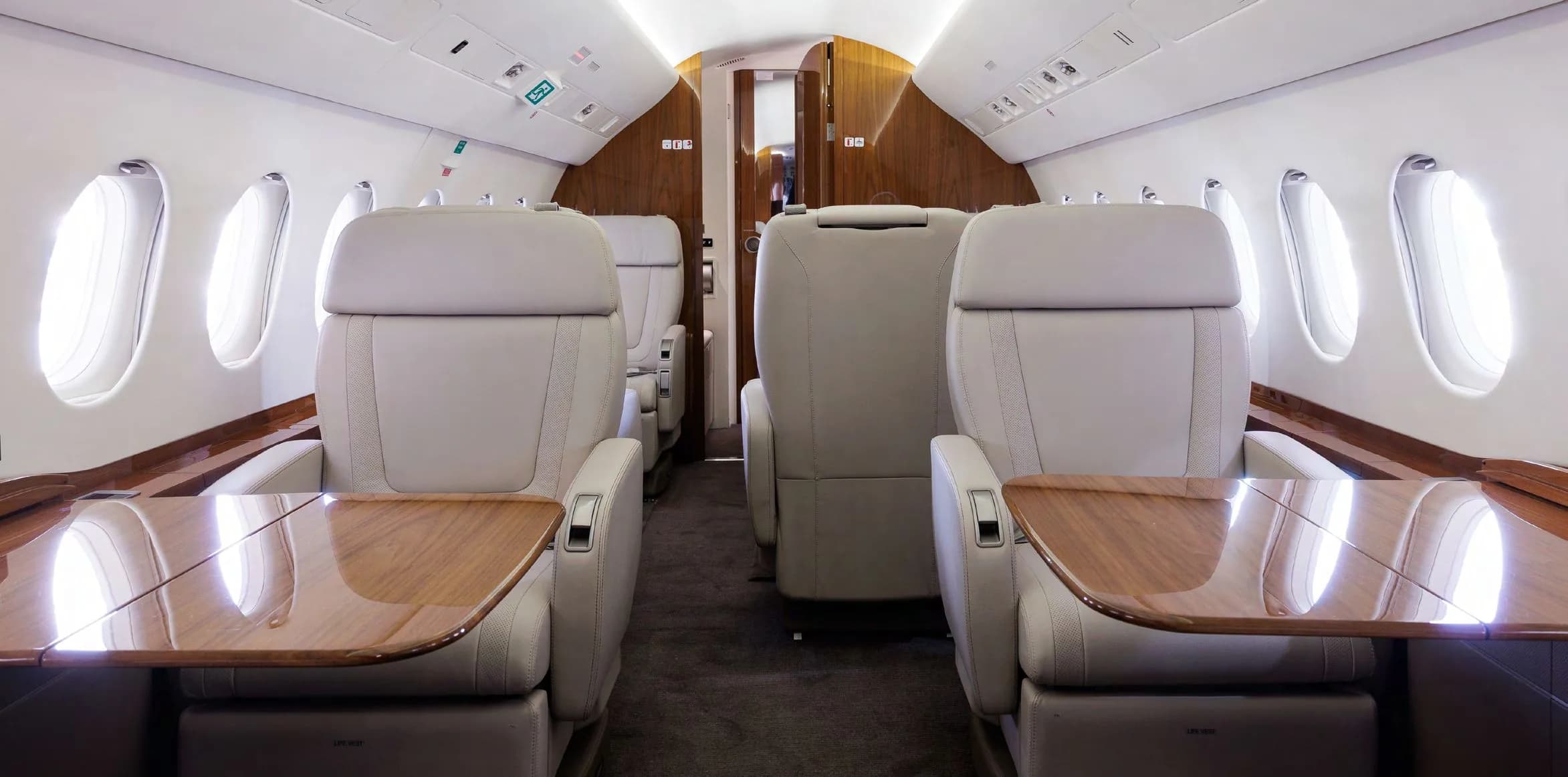 The luxurious cabin interior of a business jet with light grey leather seats and polished wooden tables.