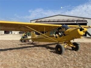 A yellow Cubcrafters aircraft is parked in front of a hangar.
