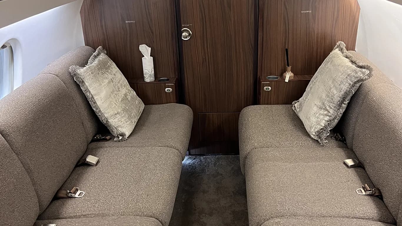 Two grey upholstered divan seats with decorative pillows face each other in the aircraft cabin.