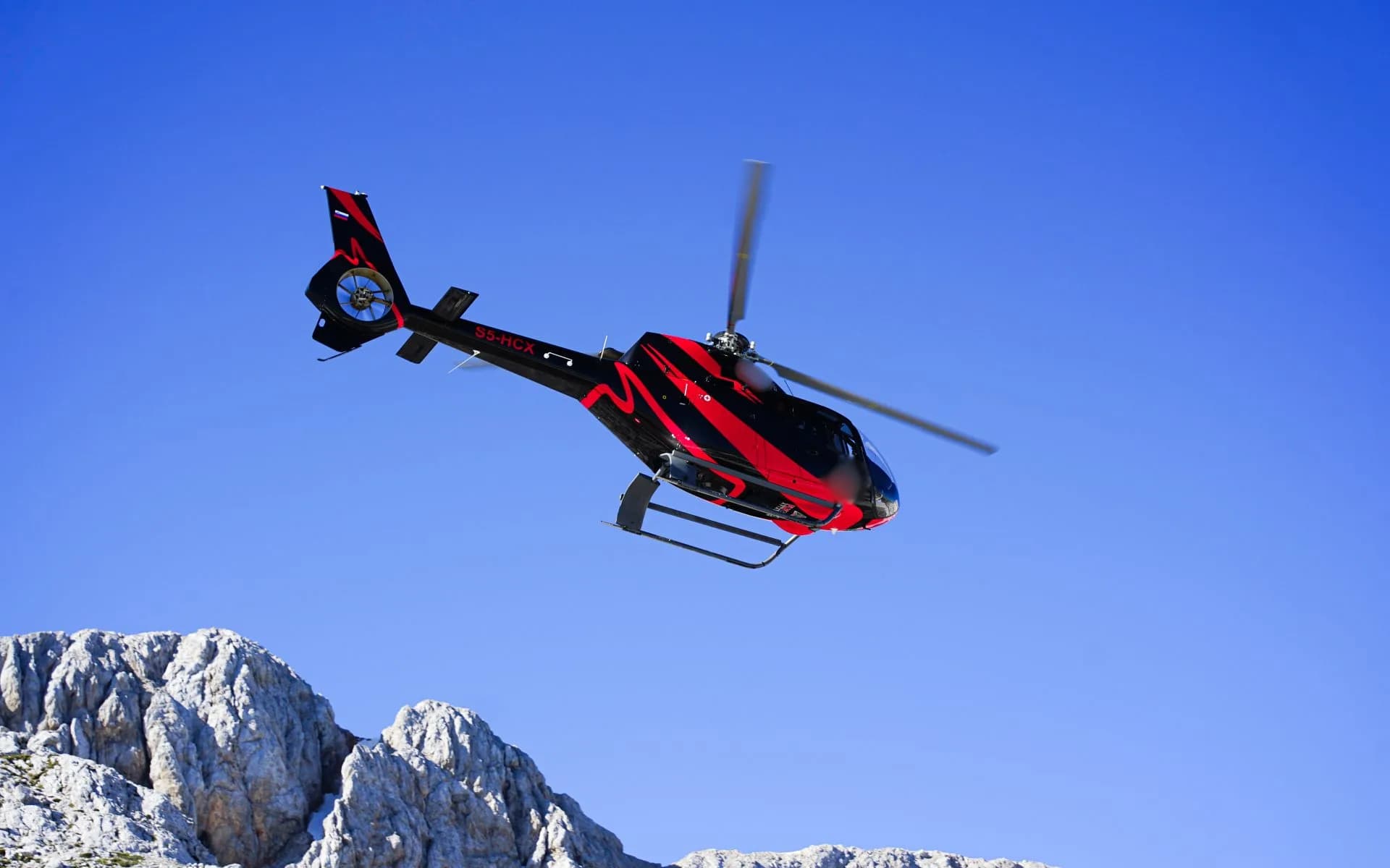 The S5-HCX helicopter is shown in flight against a blue sky, with mountains in the background.