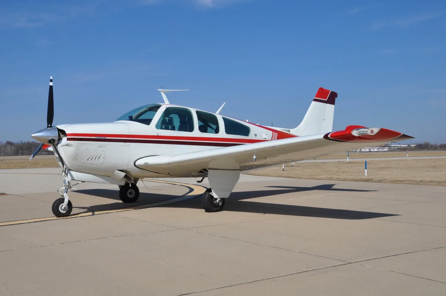 A white single-engine propeller aircraft with red and brown stripes parked on an airfield under a clear blue sky.