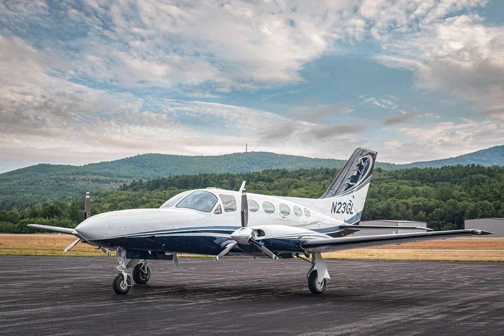 A white and blue twin-engine Cessna 414 Chancellor aircraft, tail number N23GL, parked on a tarmac with mountains in the background under a cloudy sky.