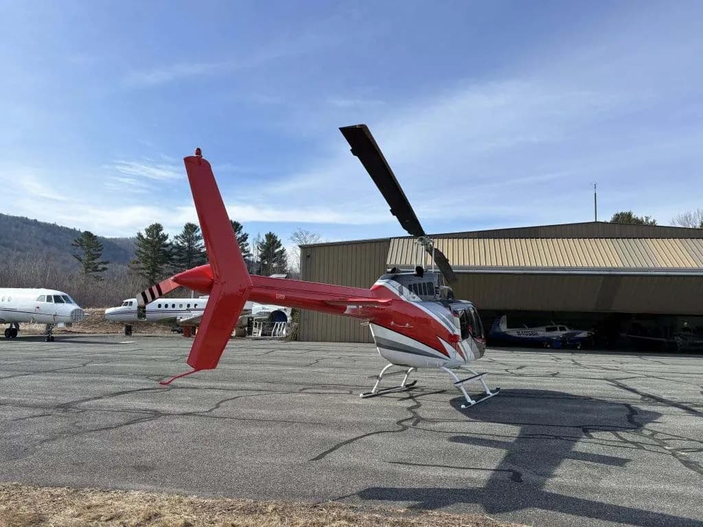 A red, black, and white helicopter parked on an airfield next to a hangar and other aircraft.