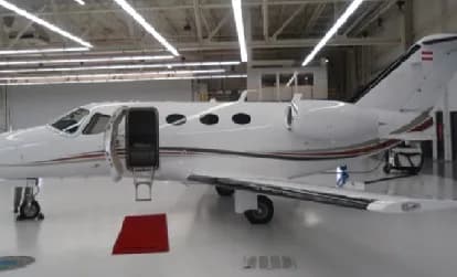 A white business jet with a red and black stripe, parked inside a hangar with its main door open.