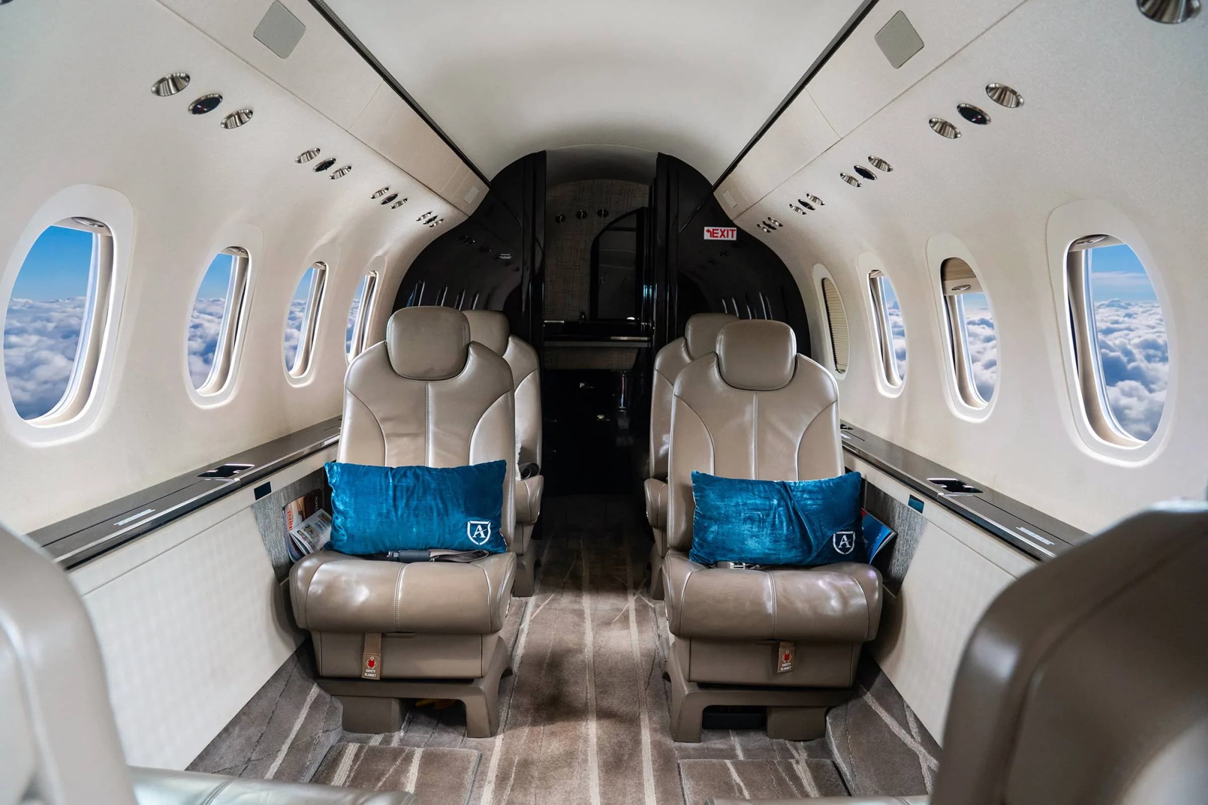 The interior of a private jet cabin with two tan leather seats, blue pillows, and windows showing clouds.