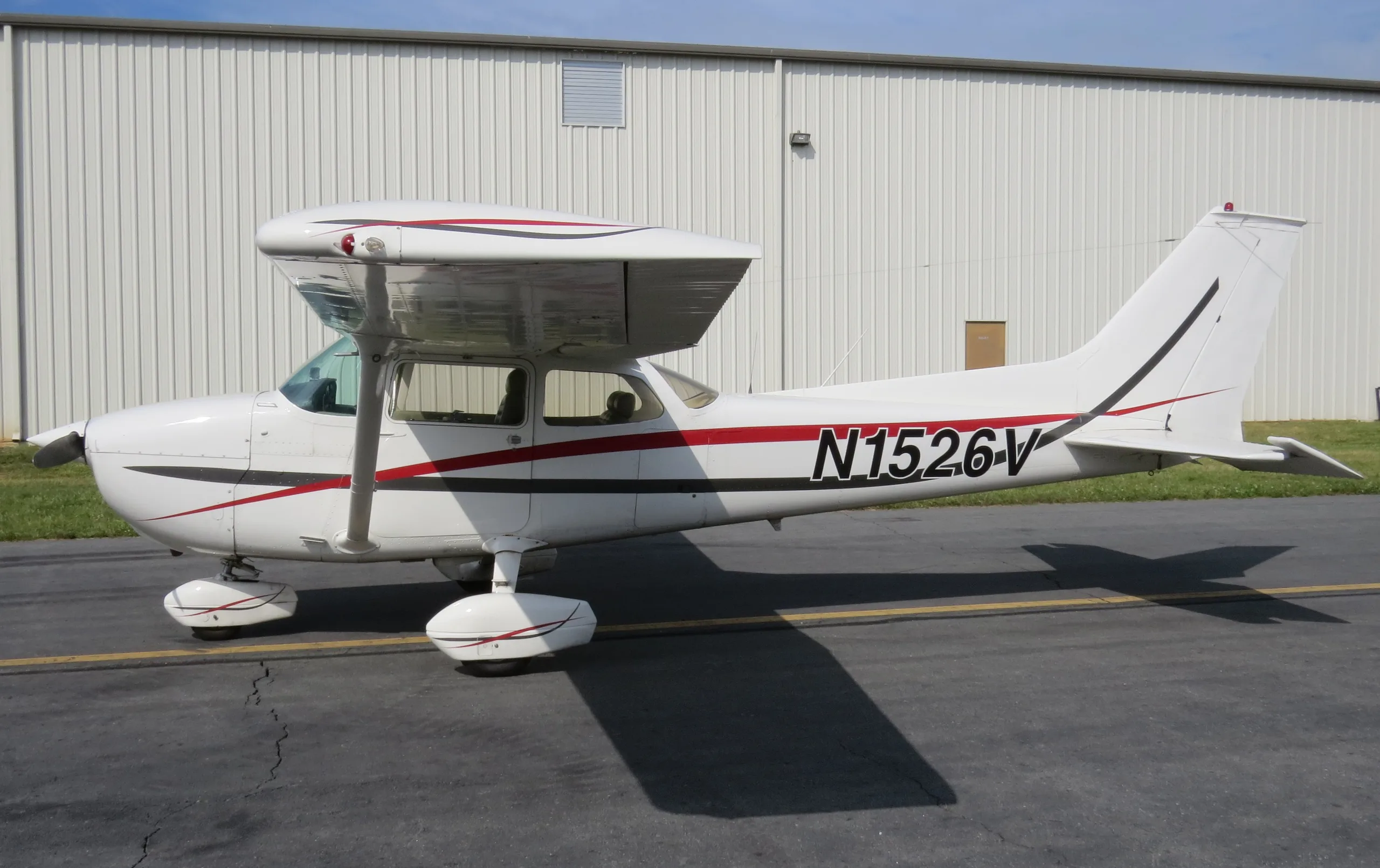 A white Cessna aircraft with red and black stripes, registration N1526V, parked on an asphalt tarmac next to a white hangar.