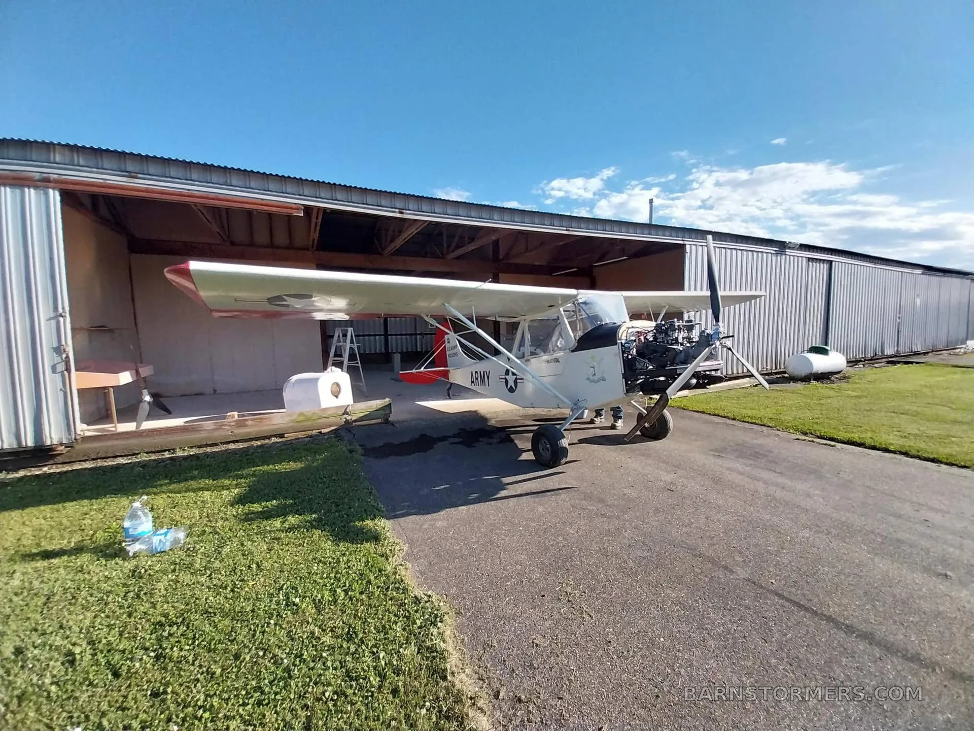 A white experimental aircraft with 'ARMY' markings and an exposed engine is parked on an asphalt ramp in front of an open hangar.