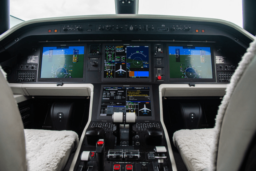 The cockpit of an Embraer Legacy 600, showing multiple digital displays, controls, and two sheepskin-covered pilot seats.