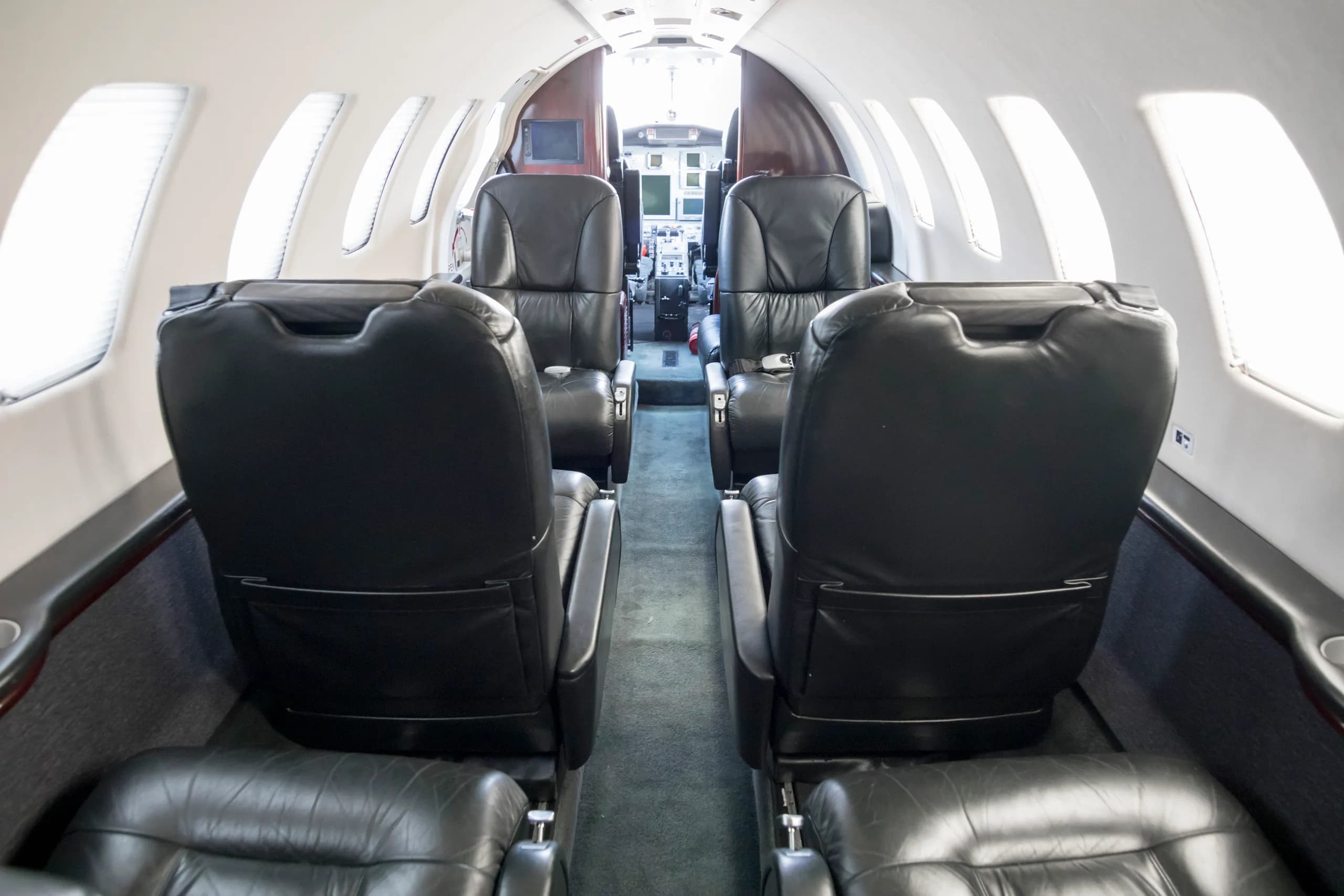 The interior cabin of a private jet, showing rows of black leather seats looking forward towards the cockpit.
