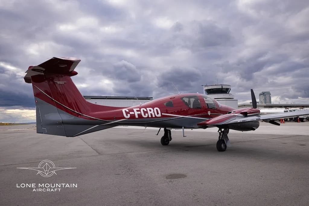 A red and grey Diamond DA62 aircraft with registration C-FCRQ parked on an airfield with a control tower in the background.