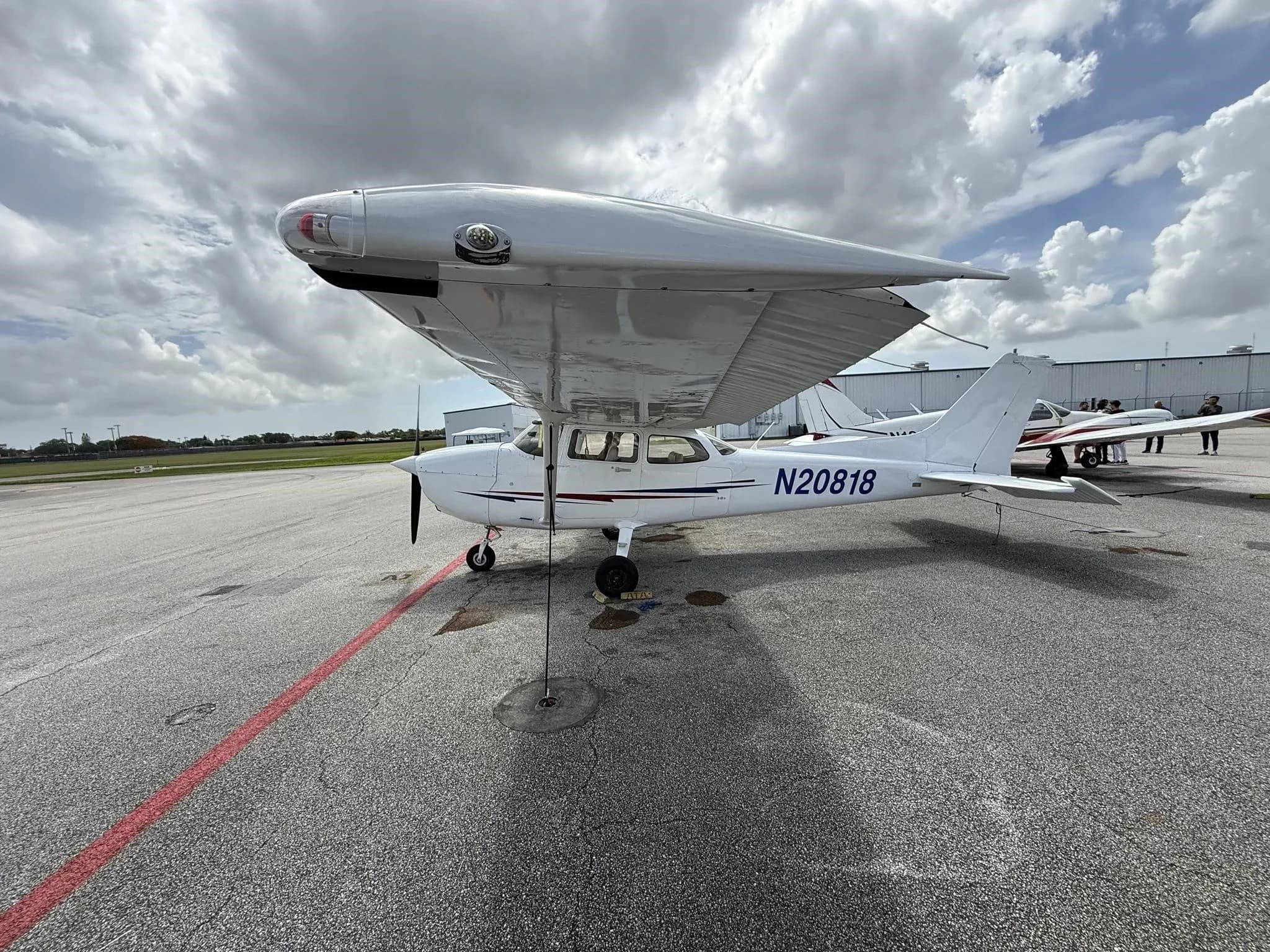 A white Cessna 172 (N20818) parked on an airport tarmac under a cloudy sky, viewed from the front-left.