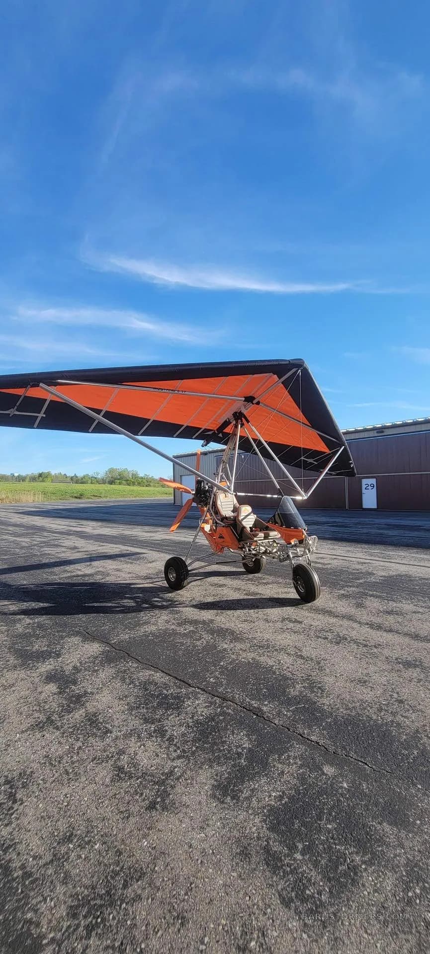 An orange and black weight-shift control ultralight aircraft parked on an airfield tarmac under a blue sky.