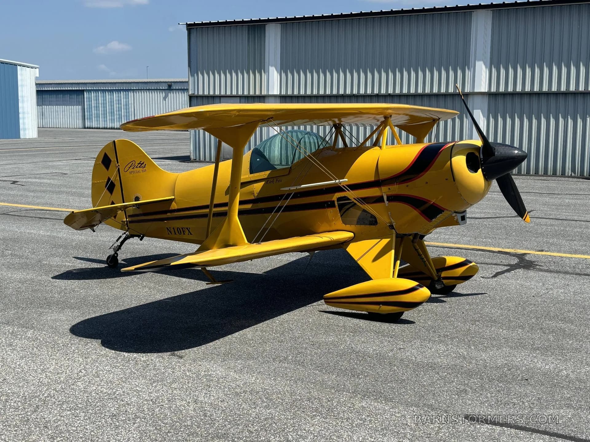 A yellow Pitts biplane with black and red stripes and registration N10FX, parked on an airfield with hangars under a clear sky.