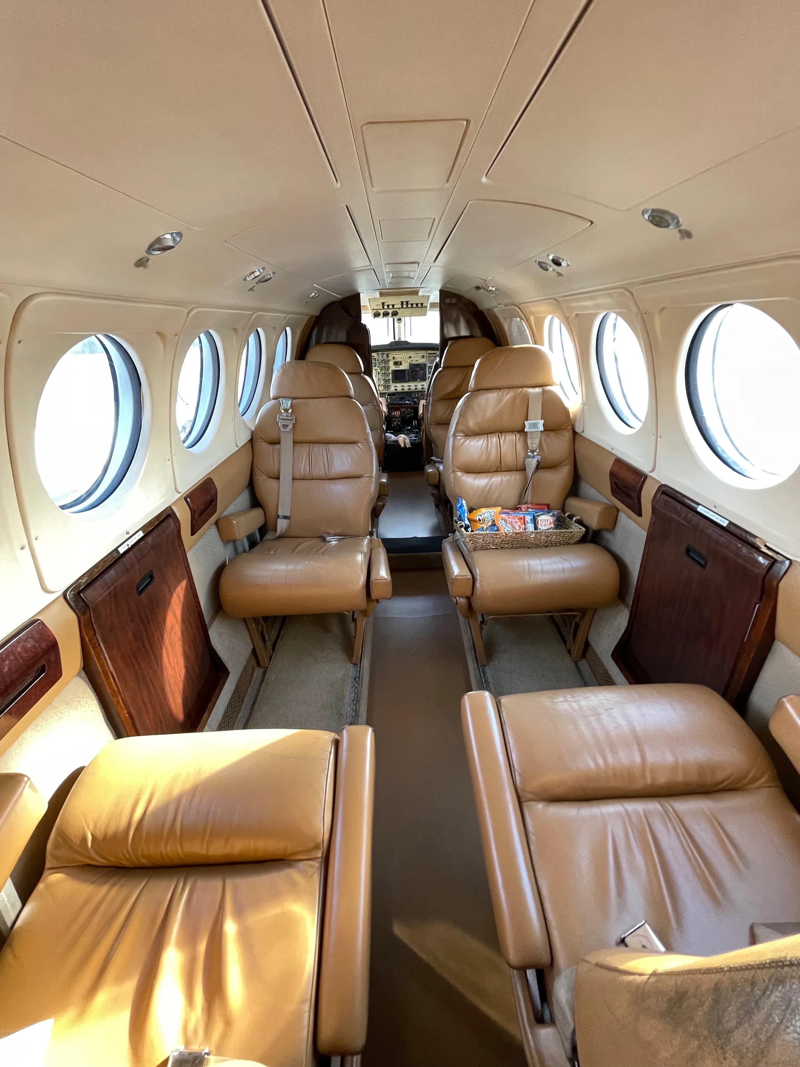 The tan leather interior cabin of a private aircraft, showing multiple rows of seats and a basket of snacks.