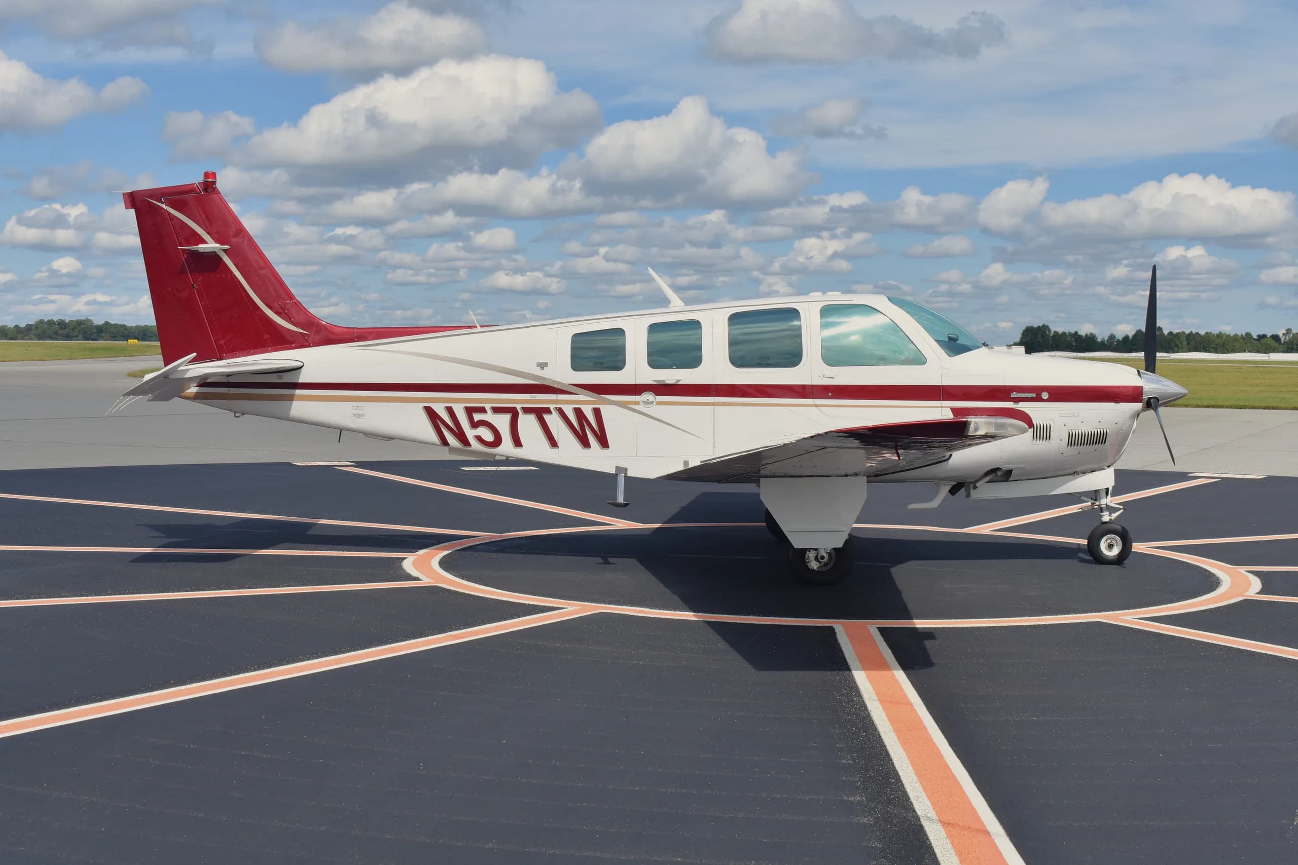 A white Beechcraft Bonanza N577TW with red and gold stripes parked on an airport tarmac under a cloudy sky.