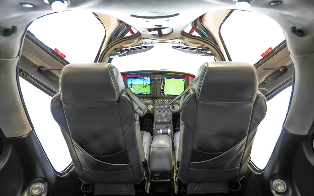 A view from the rear seats looking forward into the cockpit of a Cirrus SR22 aircraft, showing the front seats and instrument panel.