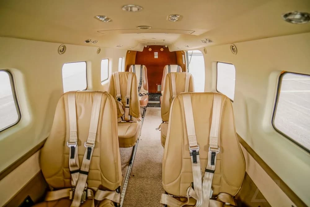 The aircraft interior cabin with tan seats and shoulder harnesses, looking towards the rear cargo area.