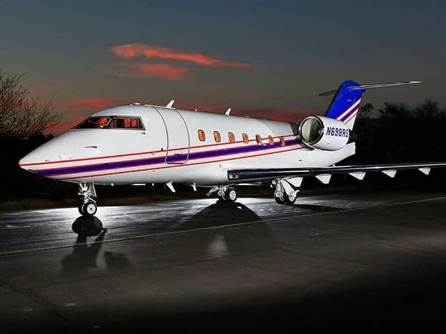 A white Bombardier Challenger business jet (N698AS) with blue and red stripes is parked on the tarmac at dusk.