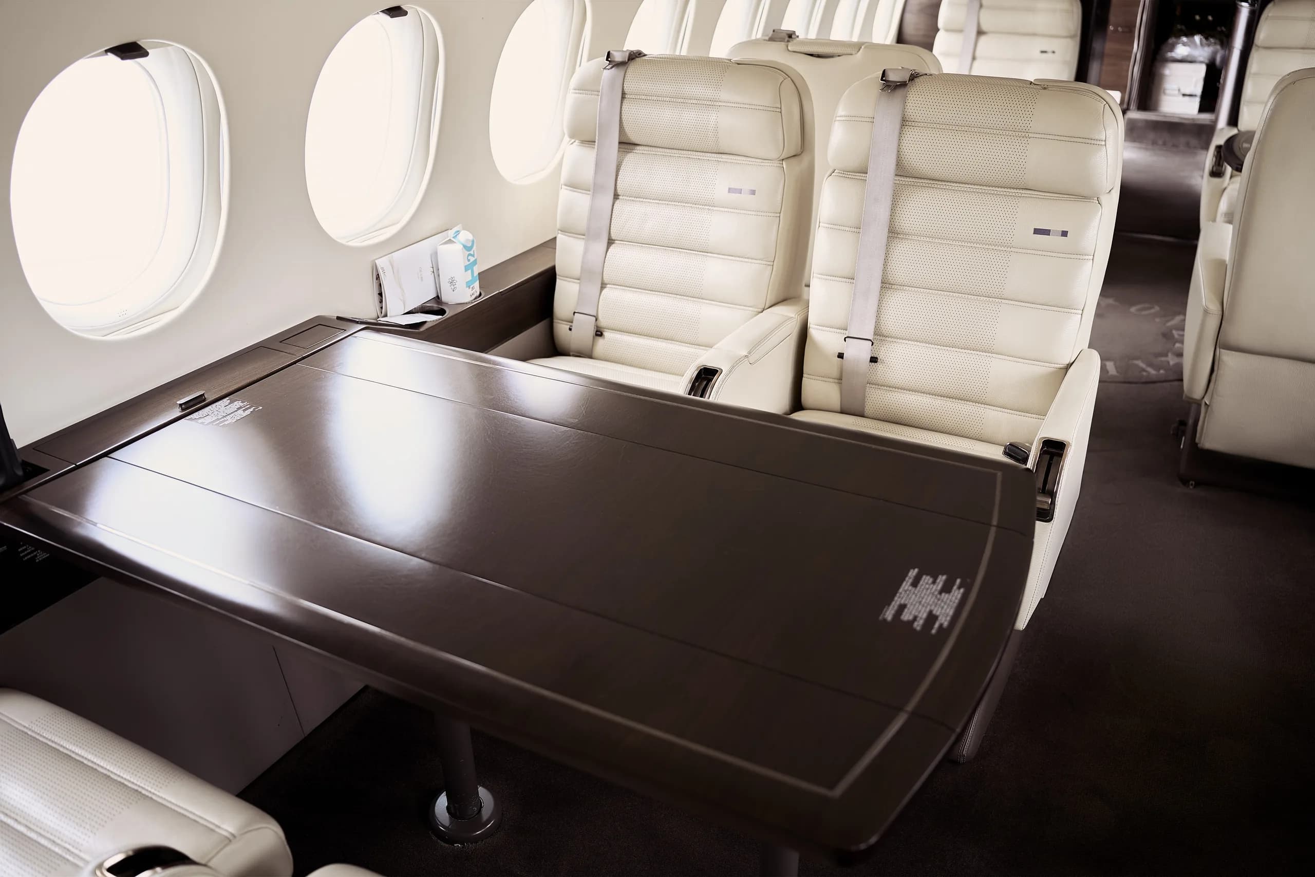 A dark wooden table with two cream leather seats next to large oval windows in a private jet cabin.
