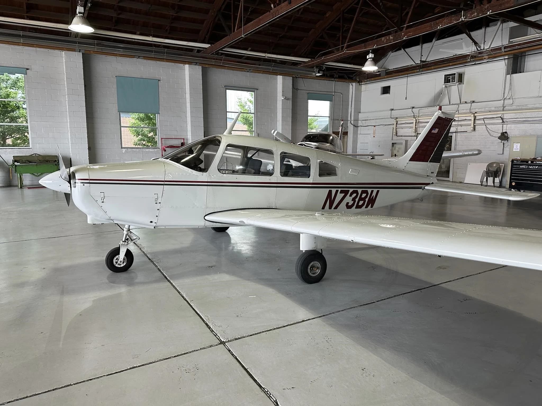 A white single-engine aircraft with maroon and black stripes, registration N73BW, viewed from the front-left in a hangar.