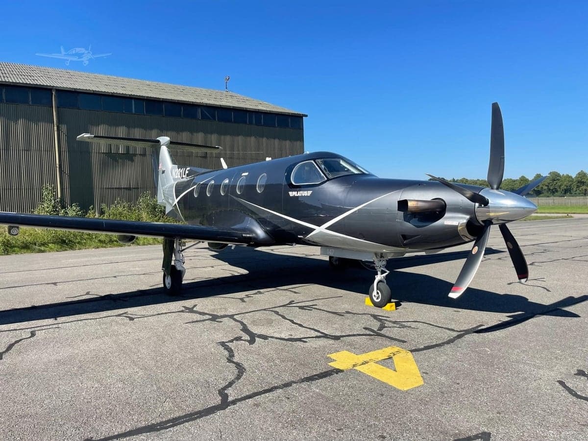 A dark grey and silver Pilatus PC-12 turboprop aircraft with white accents is parked on an asphalt tarmac next to a hangar.
