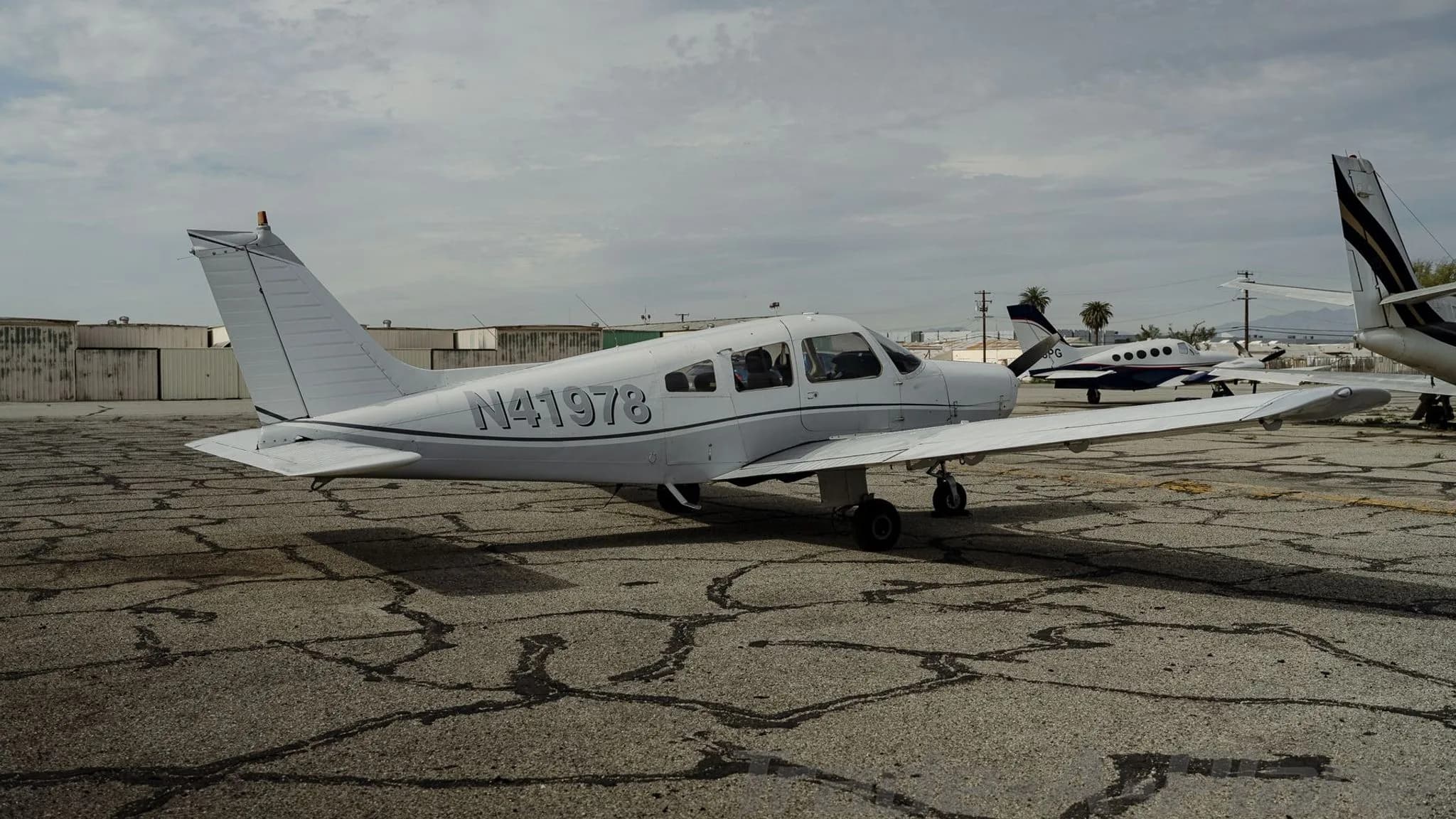 A white Piper Cherokee N41978 parked on a cracked tarmac with other aircraft and mountains in the distance.