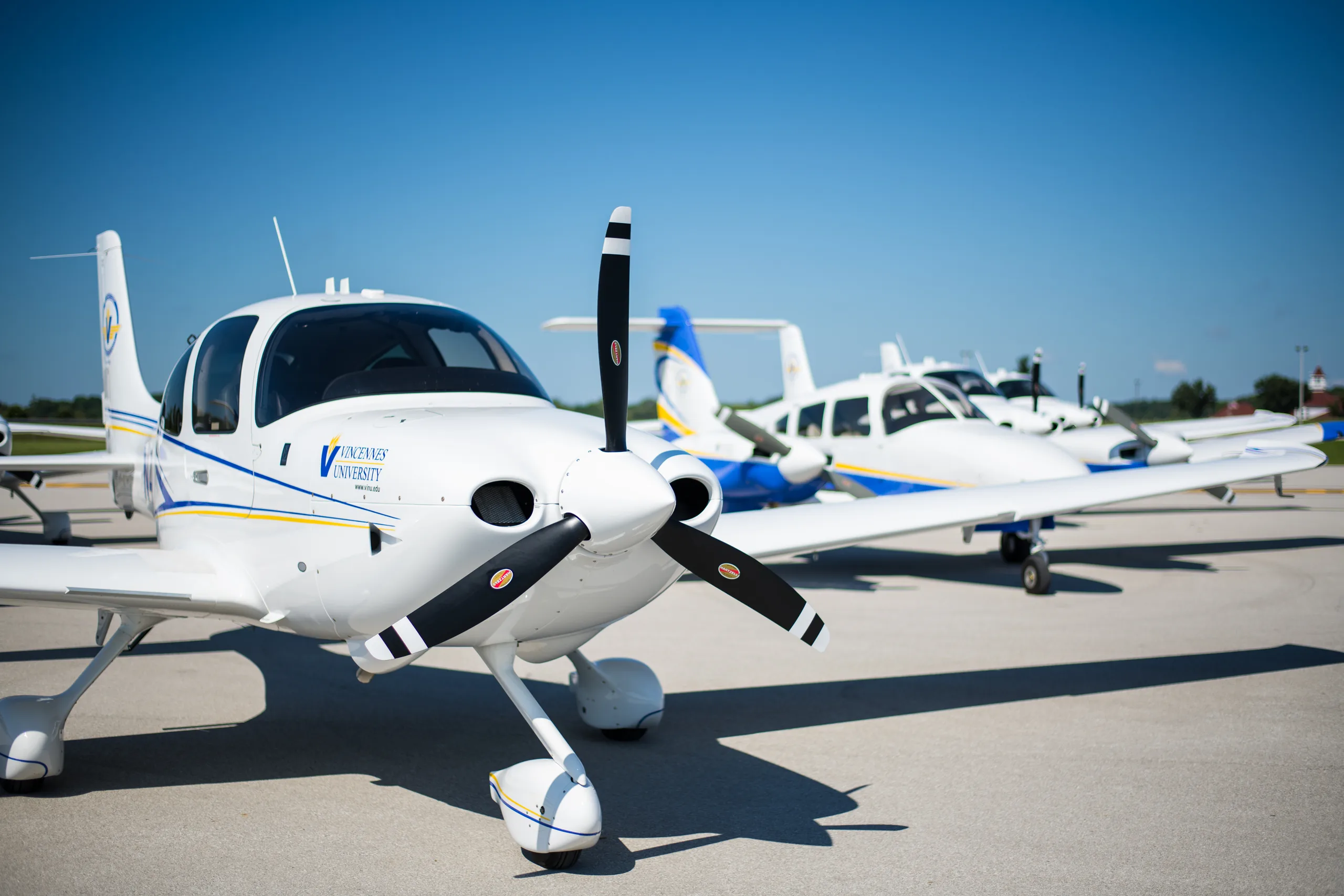 A white single-engine propeller aircraft with blue and yellow stripes and "Vincennes University" logo, parked on an asphalt runway with other aircraft in the background under a clear blue sky.