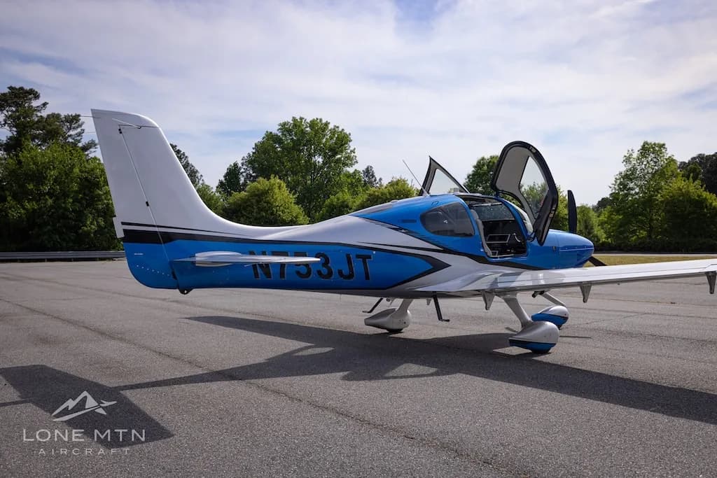 A blue and silver single-engine aircraft, N753JT, with open gull-wing doors is parked on an asphalt tarmac with trees in the background.