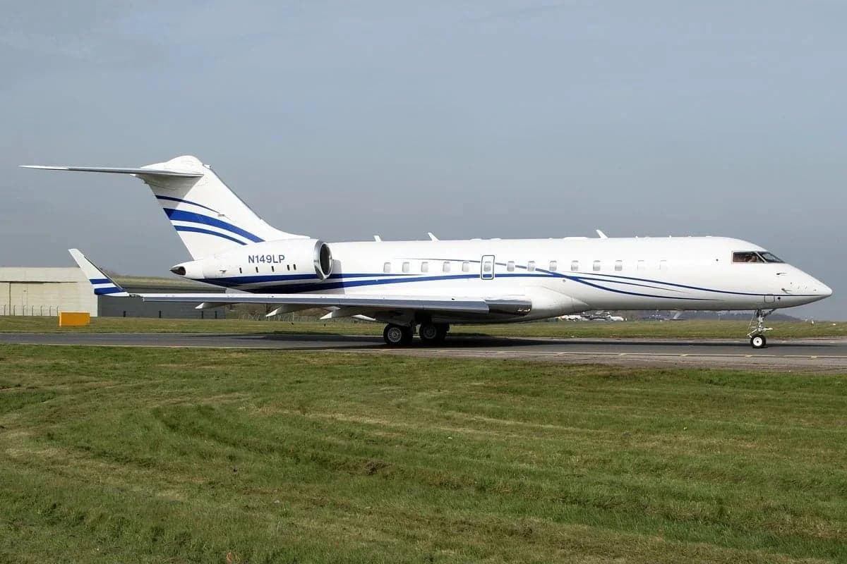 A white Bombardier Global 6000 business jet with blue stripes on its tail and fuselage, taxiing on a runway next to green grass.